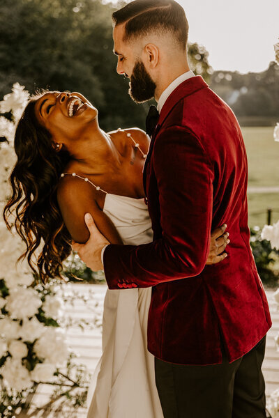 Mariée éclatant de rire dans les bras de son mari en costume de velours rouge lors de la cérémonie laïque. Moment capturé par laura, photographe de mariage en vendée.