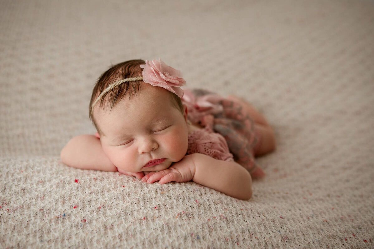 Sweet newborn girl dreams soundly, posed with hands under her chin in soft pink lace and florals.