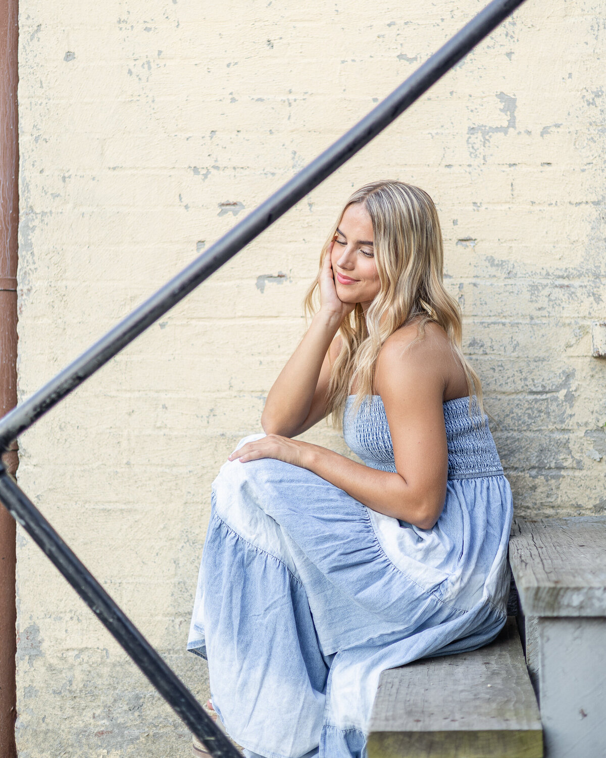 high school senior female sitting on a set of stairs with a yellow brick wall behind, with long blonde hair, wearing a blue and white sleeveless dress, smiling at the camera