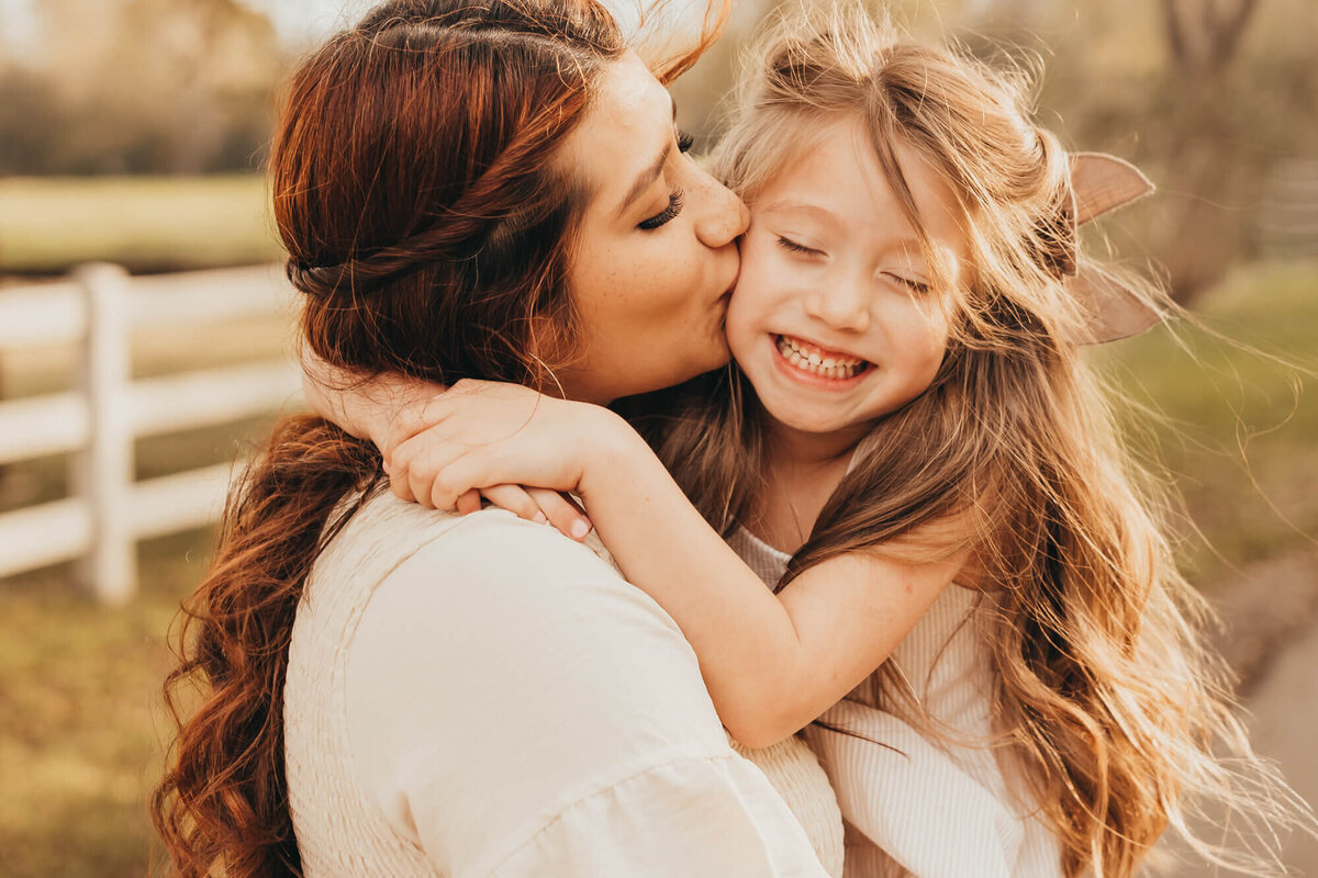 mother kisses her daughter on the cheek while she giggles.