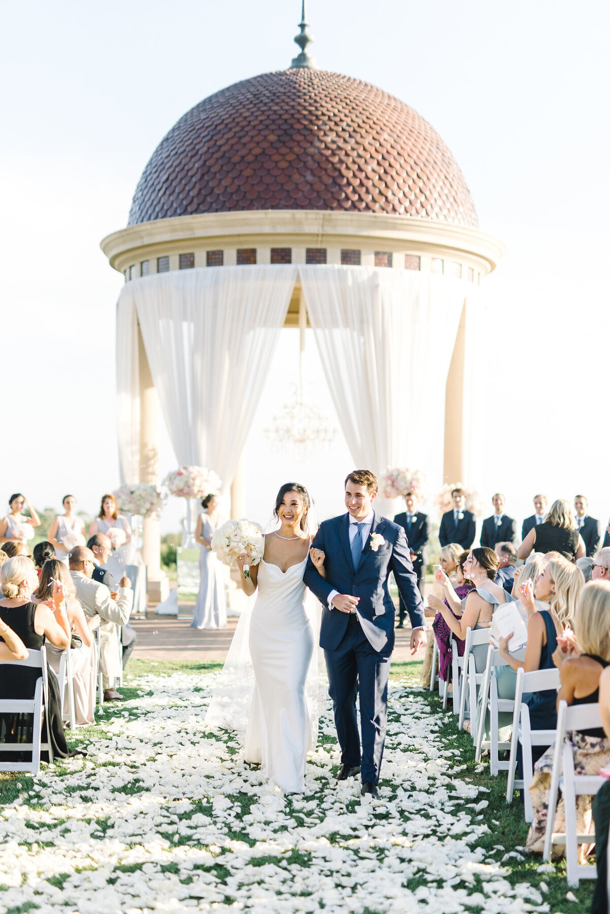 Kohnur Classic bride in a sleek satin gown with a soft glam updo and sweetheart neckline, walking recessional on a resort lawn under a grand rotunda.