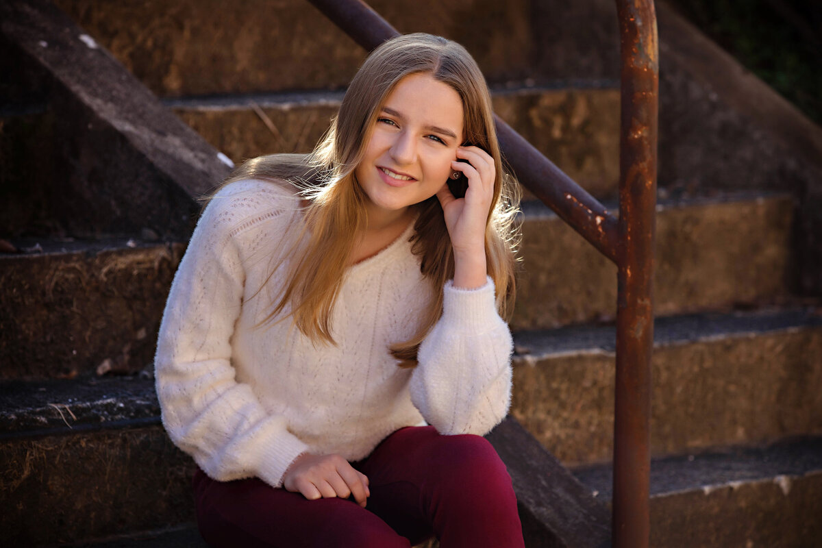 Charming HS freshman in cream sweater and cranberry pants sitting on concrete staircase with rust‑colored railings at Camp North End Charlotte NC
