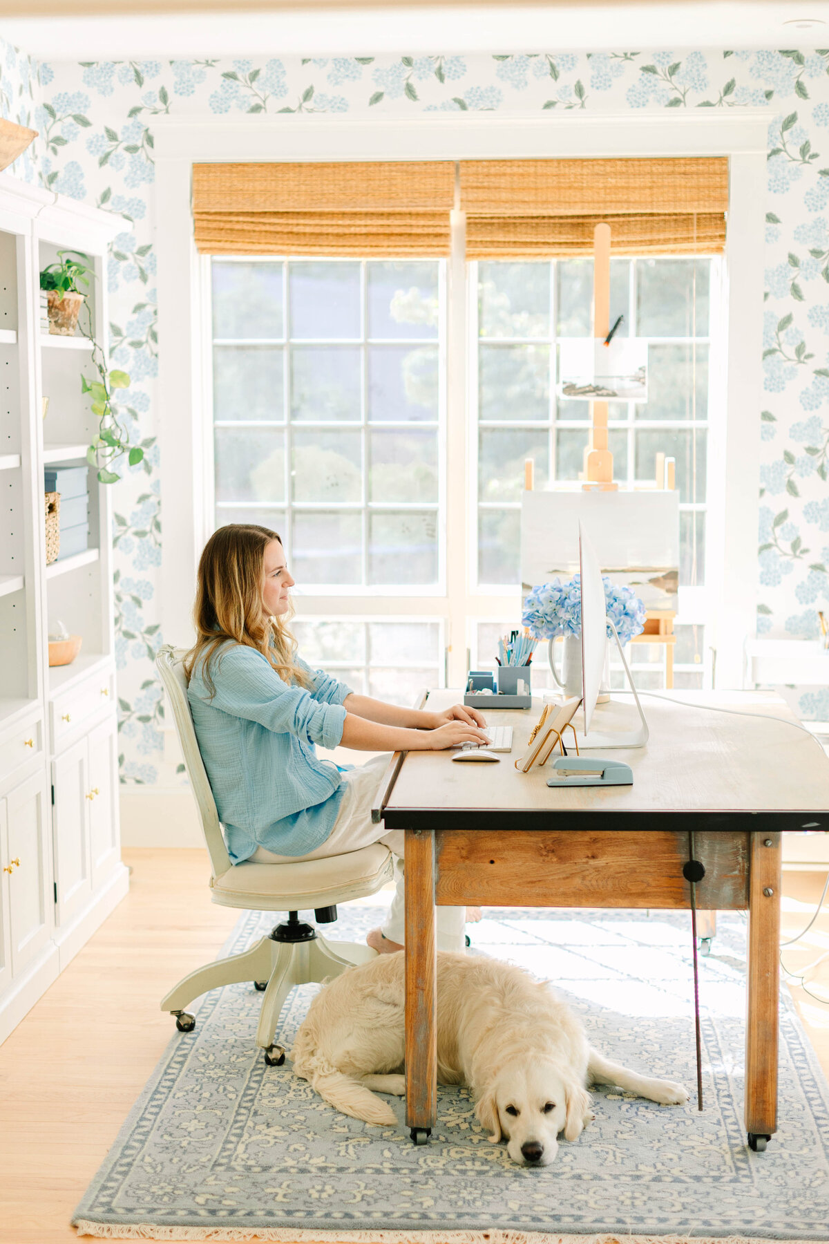 A woman in a light blue shirt sits at a wooden desk, working on a computer in a bright home office with floral wallpaper. A golden retriever rests on a blue rug beneath the desk. Natural light streams in through large windows.