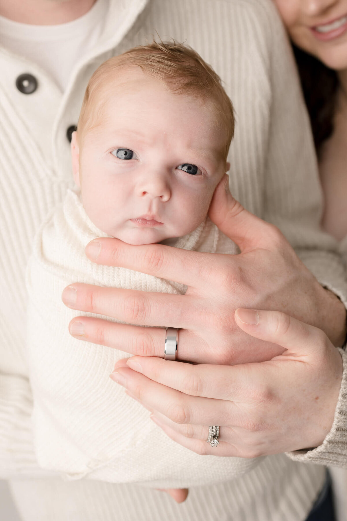 baby boy looking at camera in photo session with parent's hands holding him with their wedding rings showing