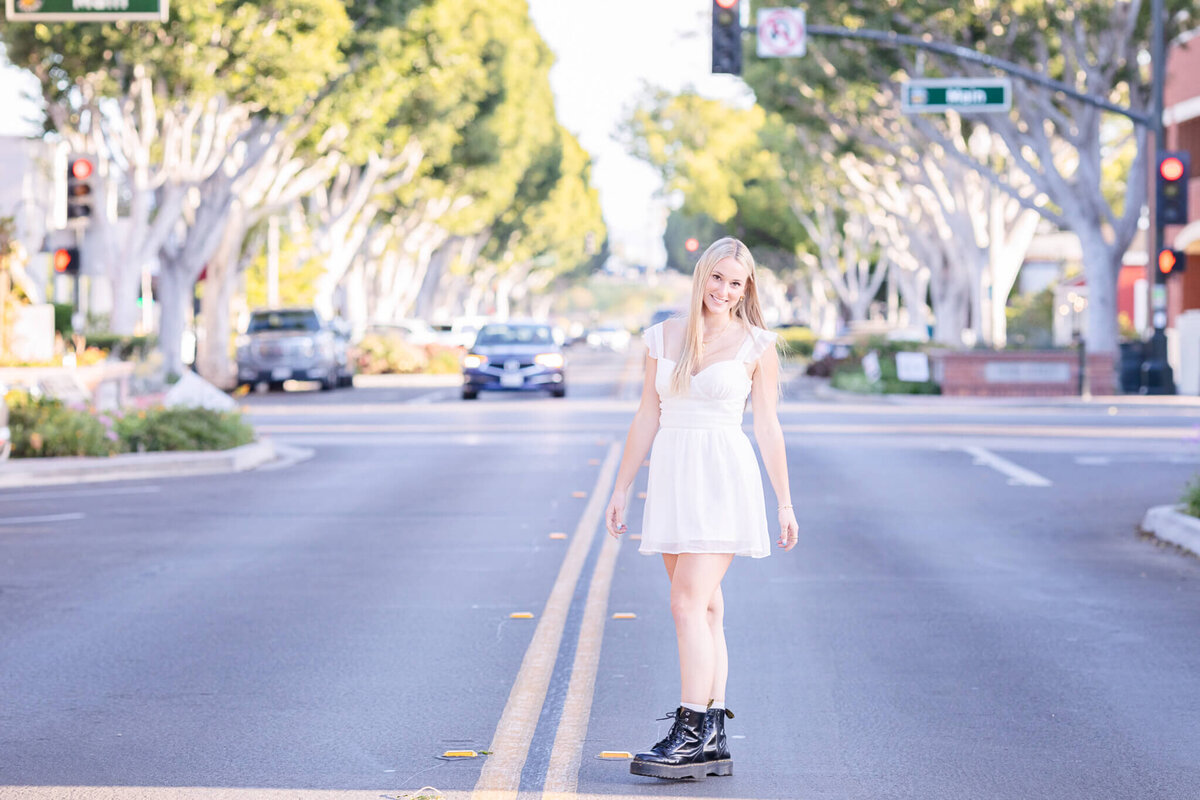 A woman in a white dress and black boots stands confidently in the middle of a tree-lined street, with cars in the background. The sunlight filters through the trees, creating a warm, inviting atmosphere.