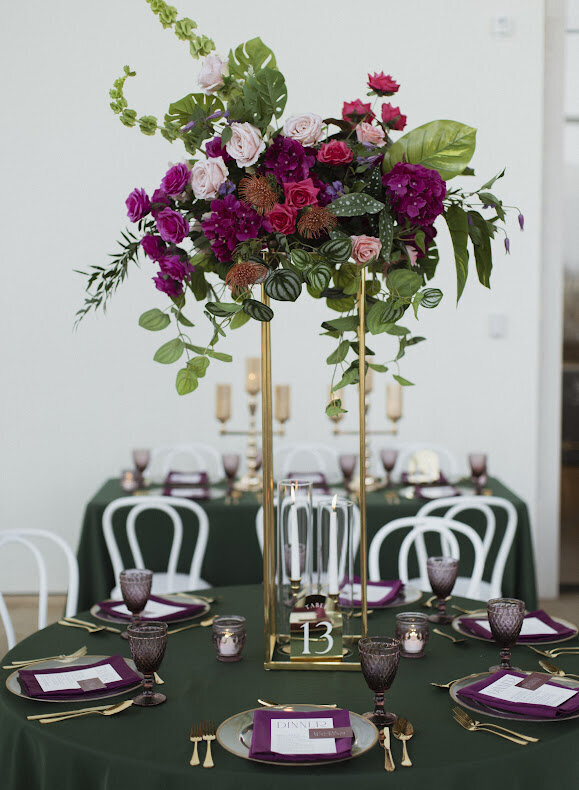 A close-up of a tropical guest table at a National Arts Centre wedding, featuring a tall fresh flower centerpiece with fuchsia blooms and tropical leaves, on top of an emerald green table linen with gold charger plates and cutlery and fuchsia napkins. and surrounded by white Bentwood chairs.