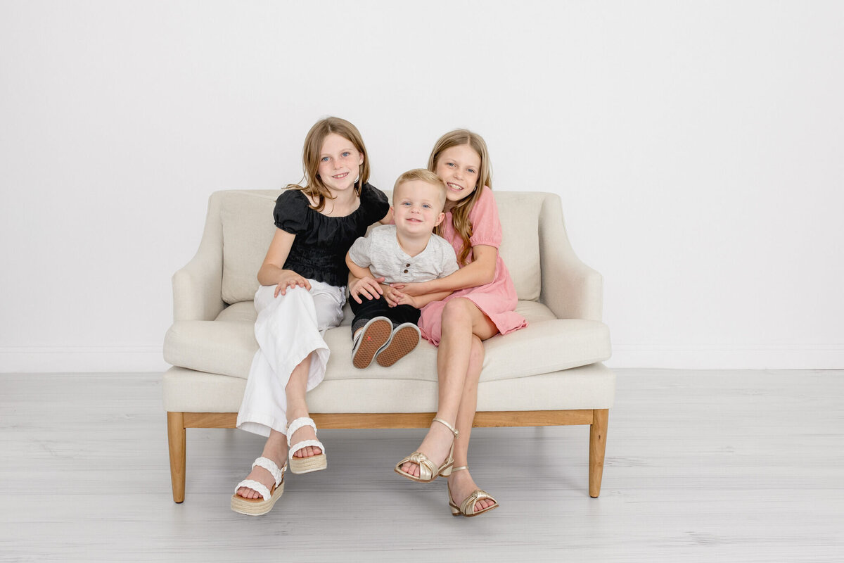 Two young girls with long blonde hair with their arms around their little brother, sitting on a couch against a white background.