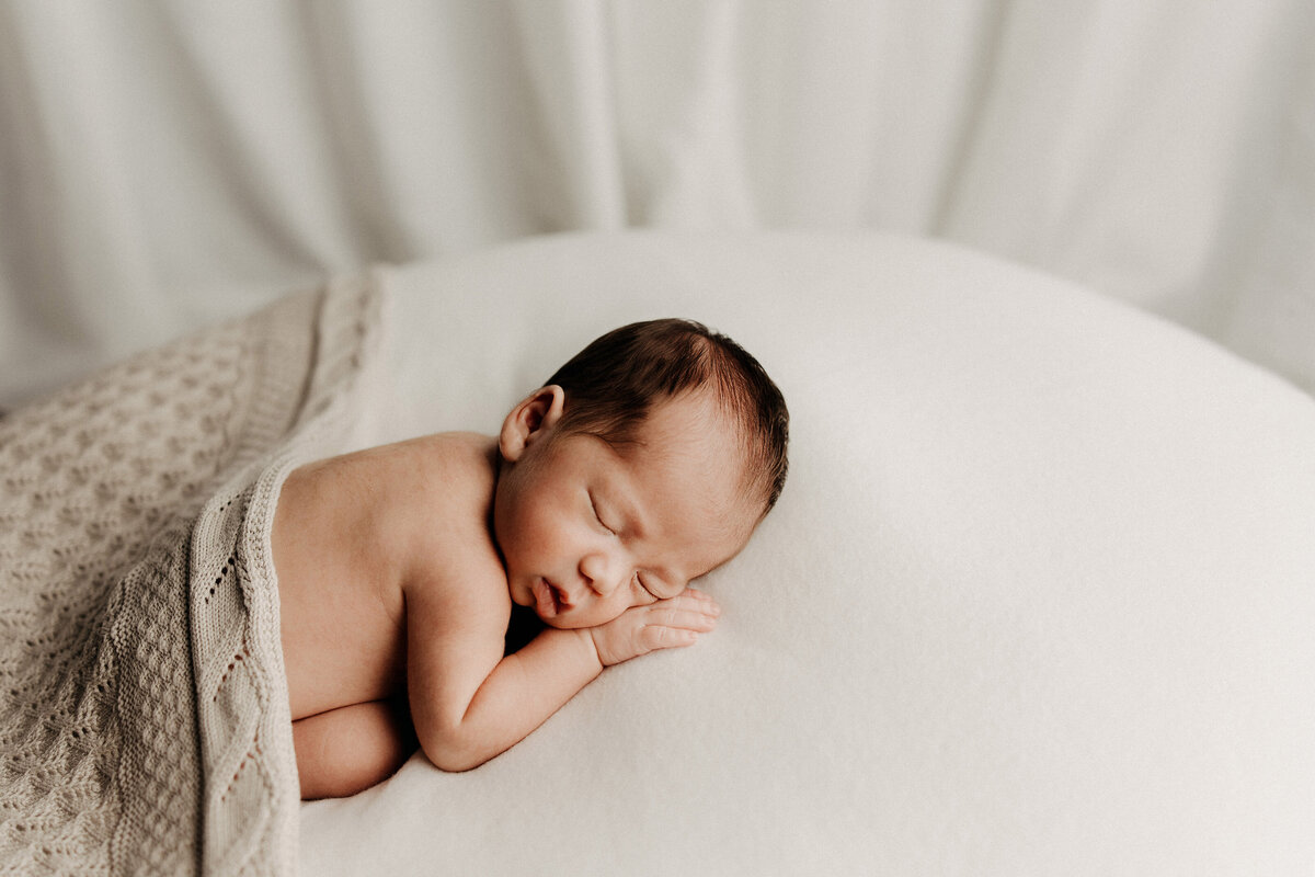Newborn baby sleeping on their side atop a soft white surface, partially wrapped in a light knit blanket.