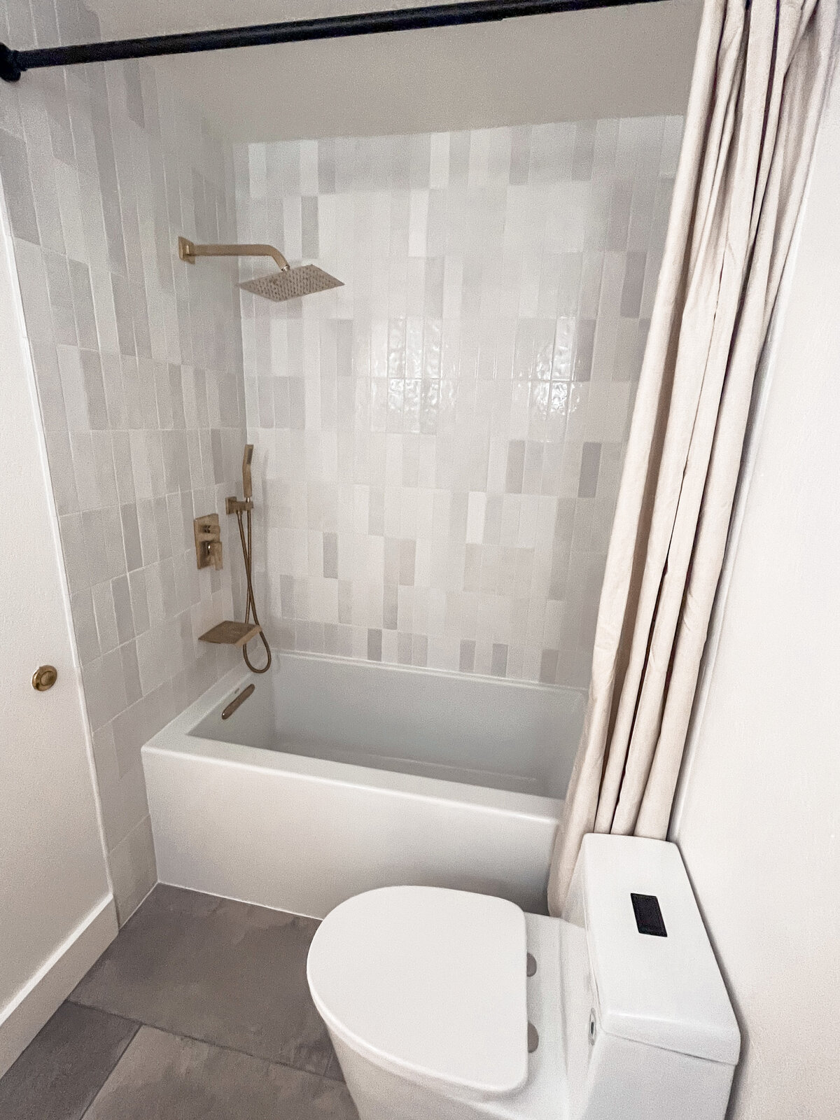 A small white bathroom with a big white soaking tub, gold hardware, and delicate grey and white vertical tiles.