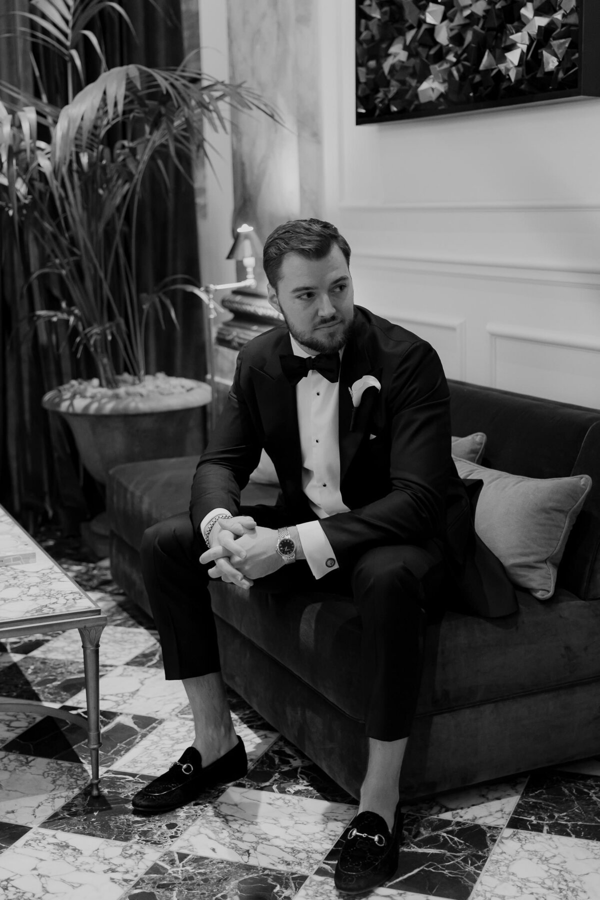 Groom in tuxedo sitting on couch in hotel looking expectantly to the side.