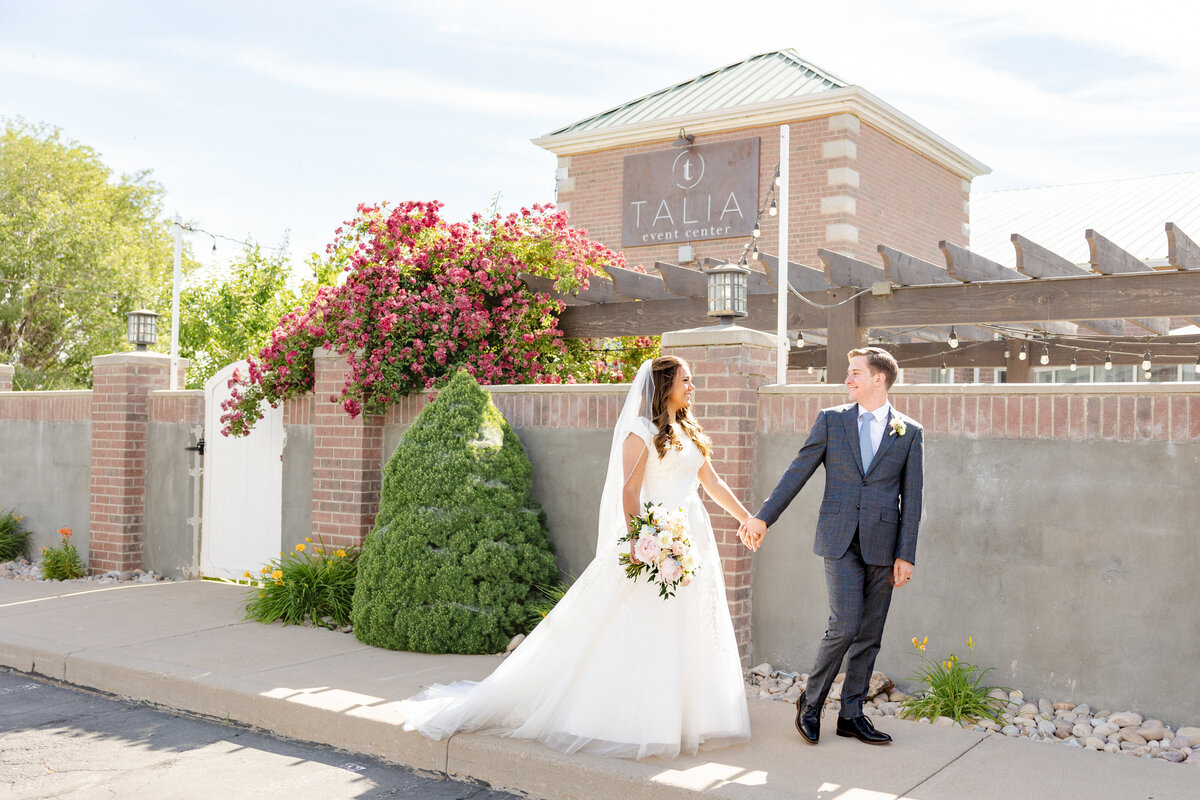 A bride and groom walk hand in hand outside the Talia Event Center, with vibrant flowers and charming brick architecture in the background.