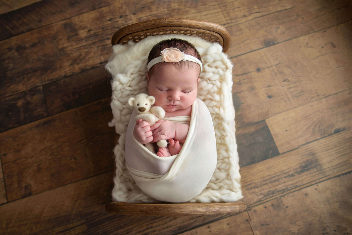 newborn baby girl wrapped in a cream colour wrap holding a cream teddybear laying in a tiny bed.