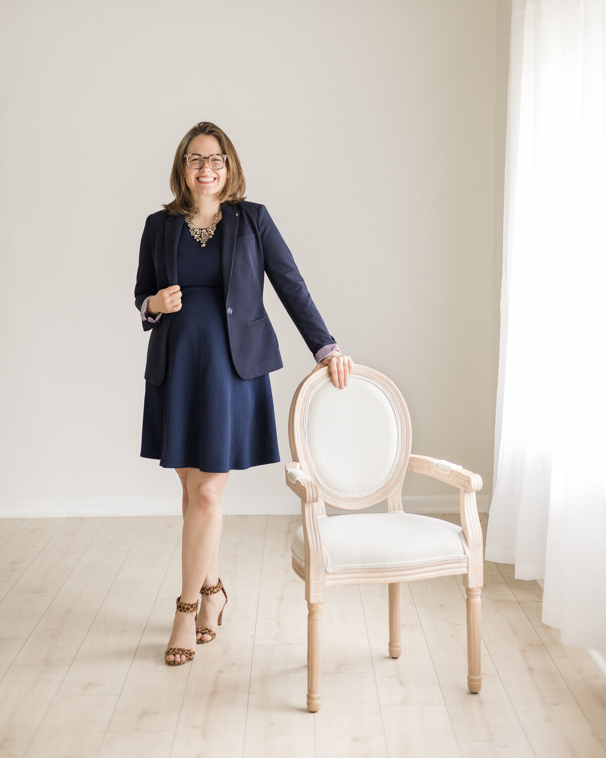 Brunette blonde pregnant female standing with her hand on a chair, wearing glasses, a dark blue dress with a long sleeve dark blue jacket, with brown and black cheetah style print heels,  smiling at the camera