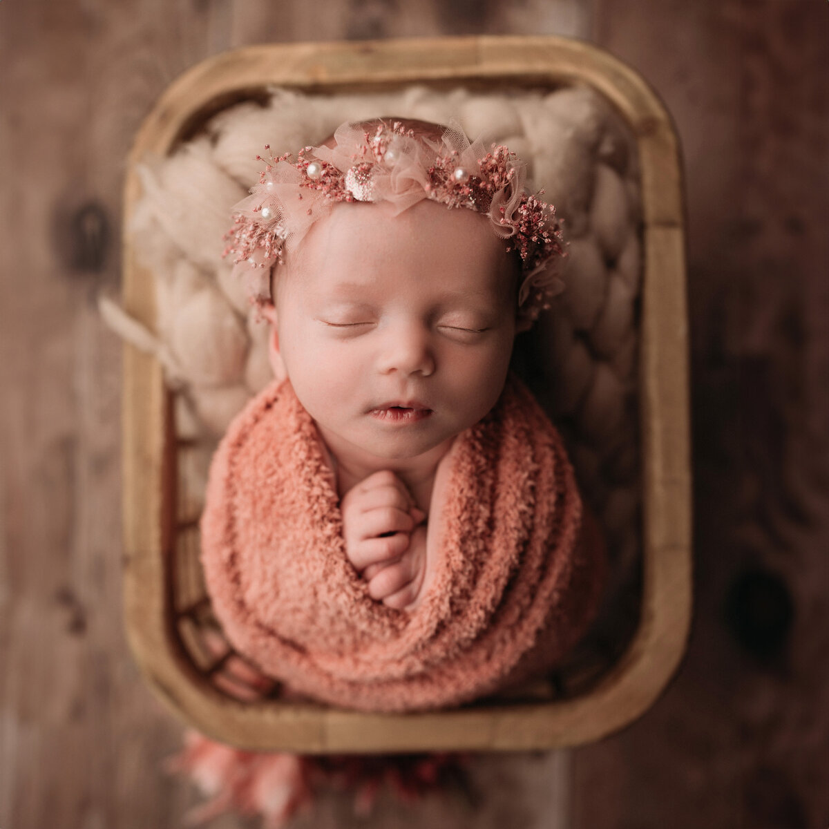 sleeping newborn baby girl wearing a rose colored flower halo and matching colored knit wrap posed inside of a rectangular wooden basket on a wooden floor