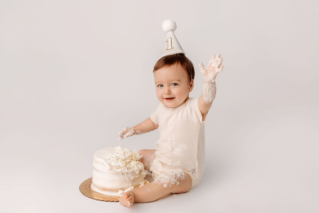 Portrait Photographers CT photograph baby celebrating first birthday with white party hat waving frosting covered hands during classic cake smash session