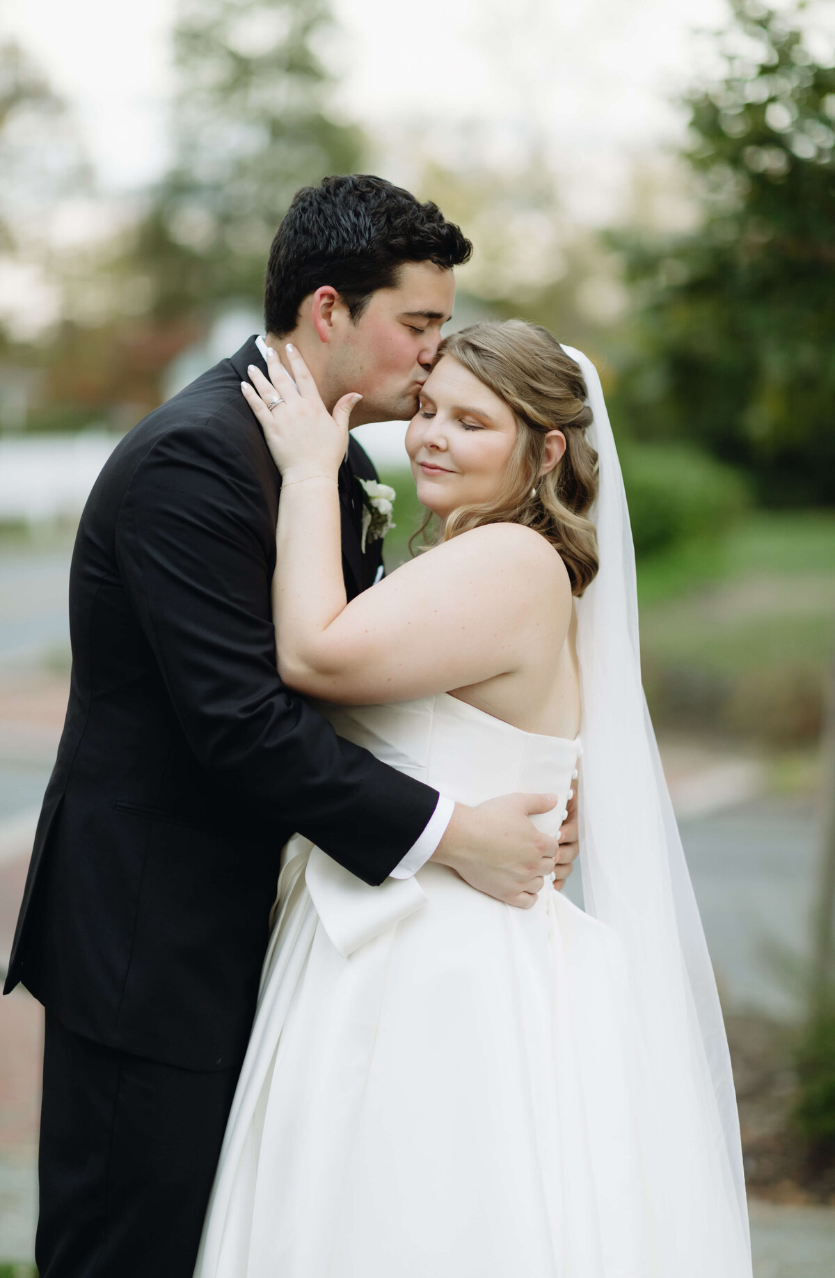 Virginia wedding photographer captures bridal photo of groom holding his bride by the waist and kissing her head as she holds her hands to his neck with a lush garden behind them