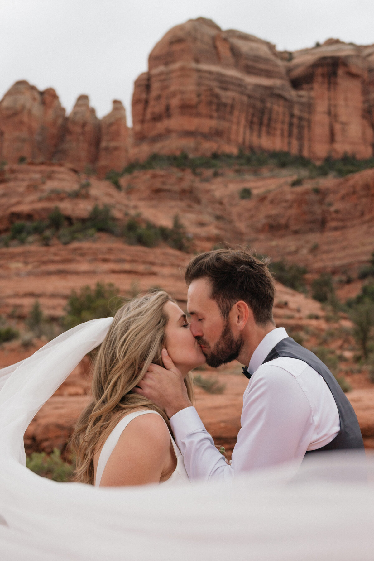Bride and groom share an intimate kiss with flowing veil and red rock formations in the background during their Cathedral Rock elopement in Sedona, Arizona.
