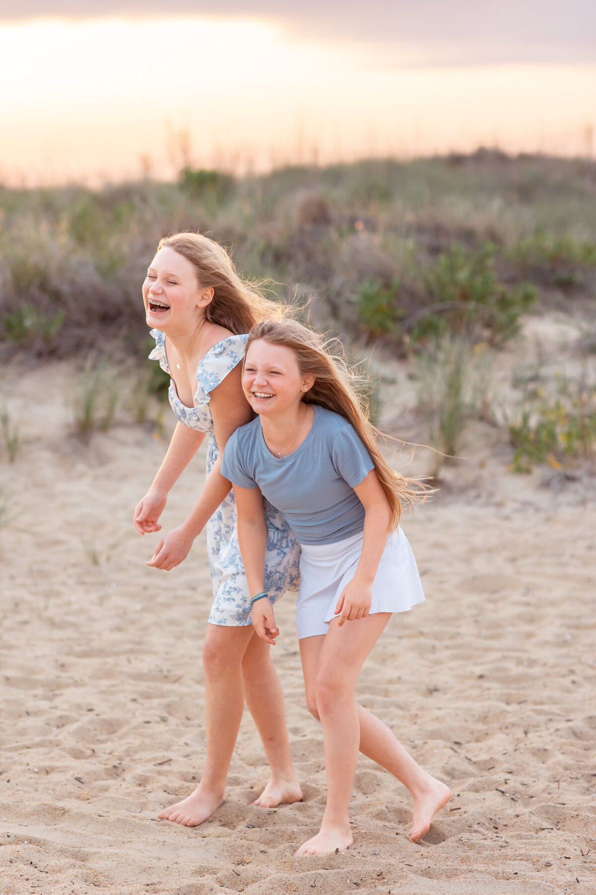 Two teenage sisters burst into laughter while running barefoot across the sandy shore, their hair caught in the breeze during a playful portrait session in Coastal Virginia with Brooke Tucker Photography