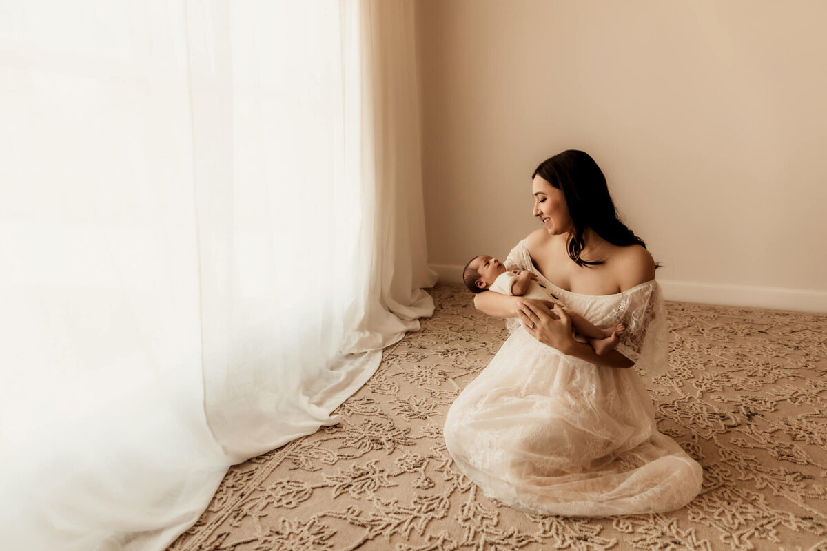 Mother kneels on rug while smiling and holding her newborn baby boy for a studio photo in OKC.