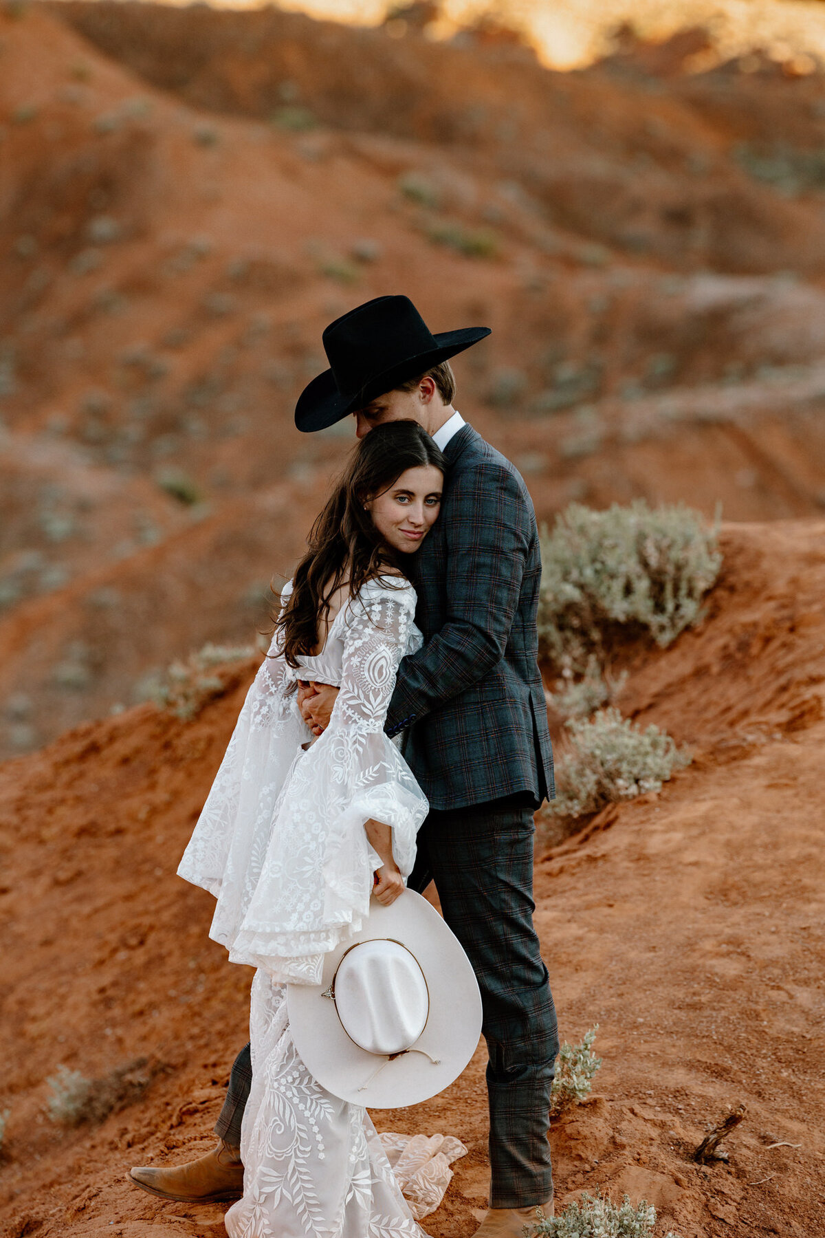 A bride looking at the camera snuggles into the chest of her cowboy hat wearing groom. 