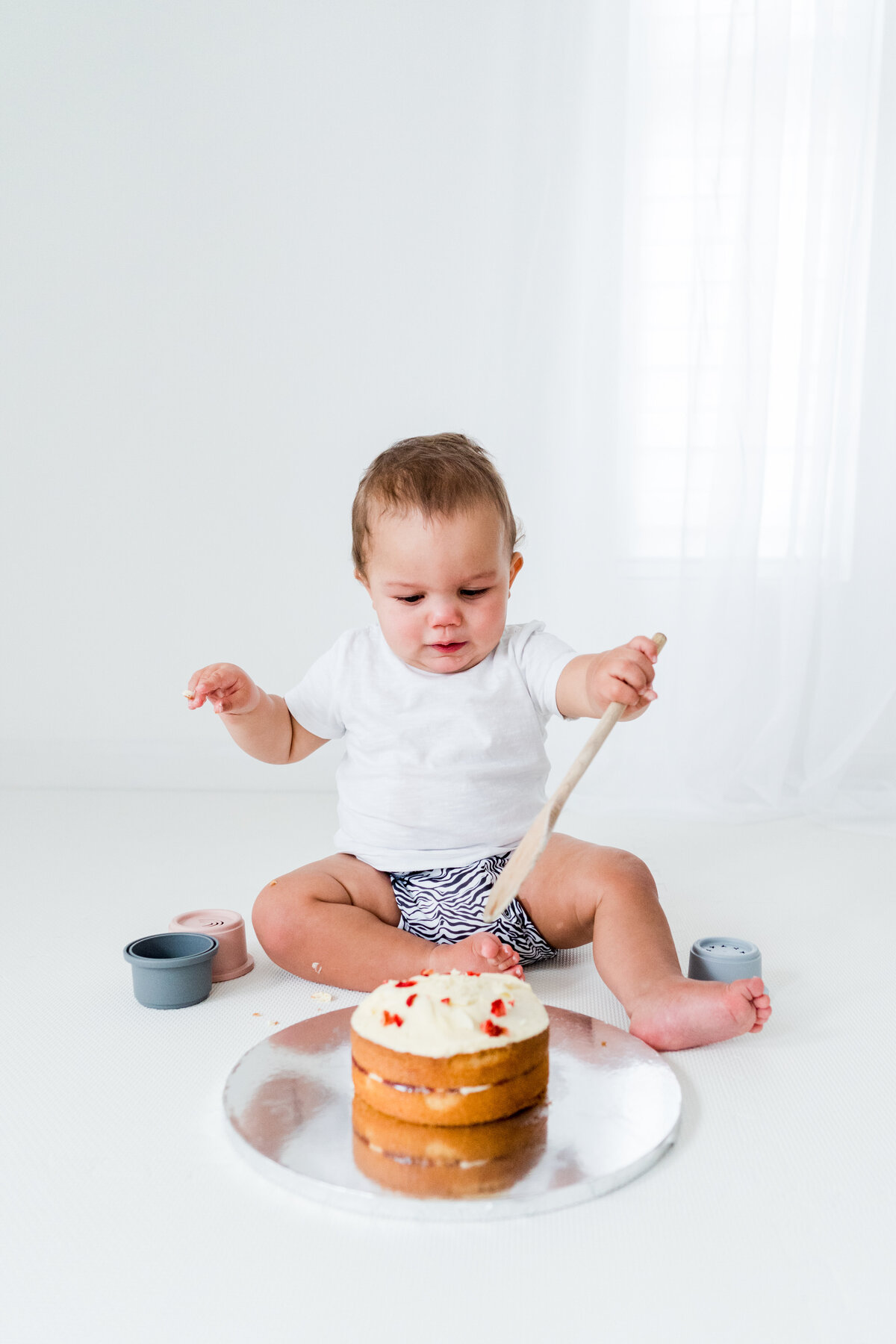 little boy uses a wooden spoon to 'smash' his cake at cake smash photo shoot