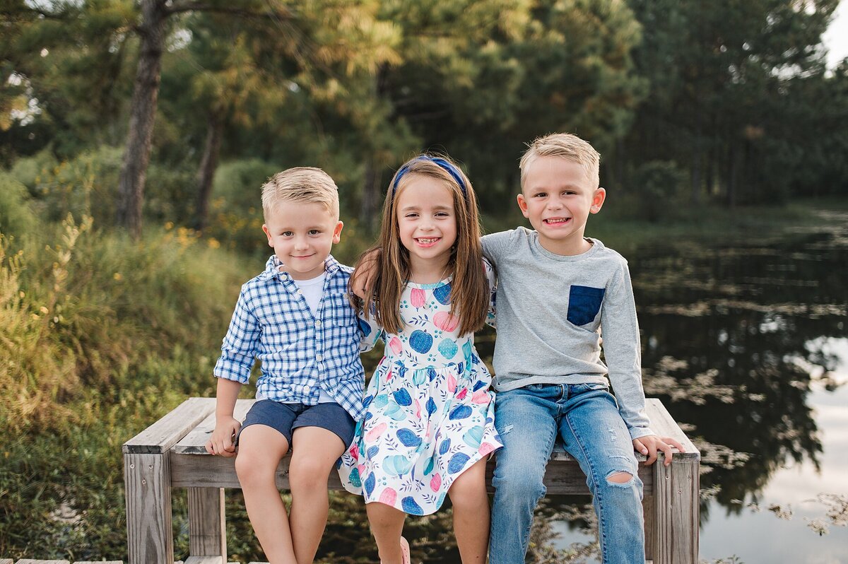 two brothers and their sister sitting on wooden bench by a pond by Cypress Family Photographer
