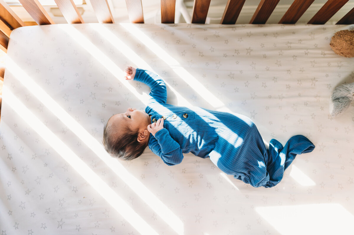 newborn baby boy in blue lying in crib with window light streaming in
