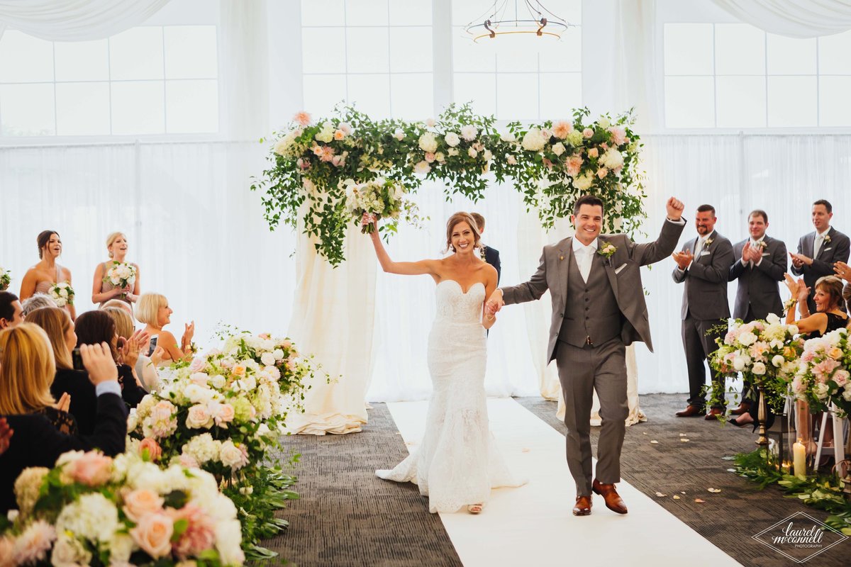 wedding couple just married walking down aisle in front of large greenery and flower chuppah in white tent