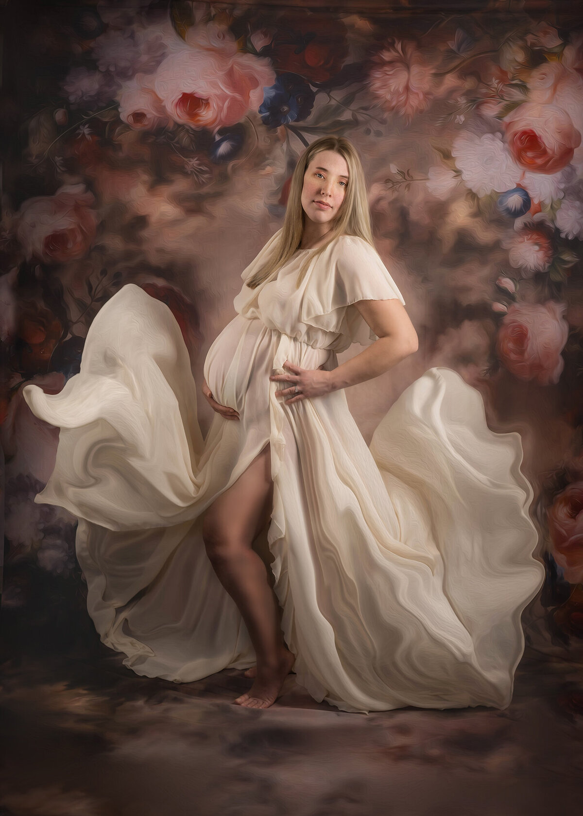A mom to be in a flowing white maternity dress stands in a studio with a carnation floral backdrop for some Iowa maternity photography