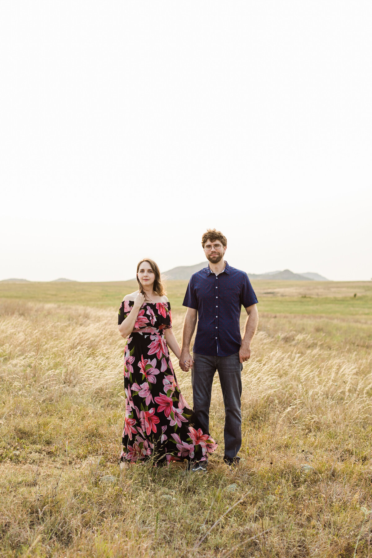 A couple holding hands in a field during their anniversary photo session in the Wichita Mountains in Oklahoma. The woman on the right is holding her hair with her right hand and is wearing a long black dress covered in large colorful flowers. The man on the right is wearing a blue dress shirt and dark means. Some of the Wichita Mountains can be seen far off in the background.