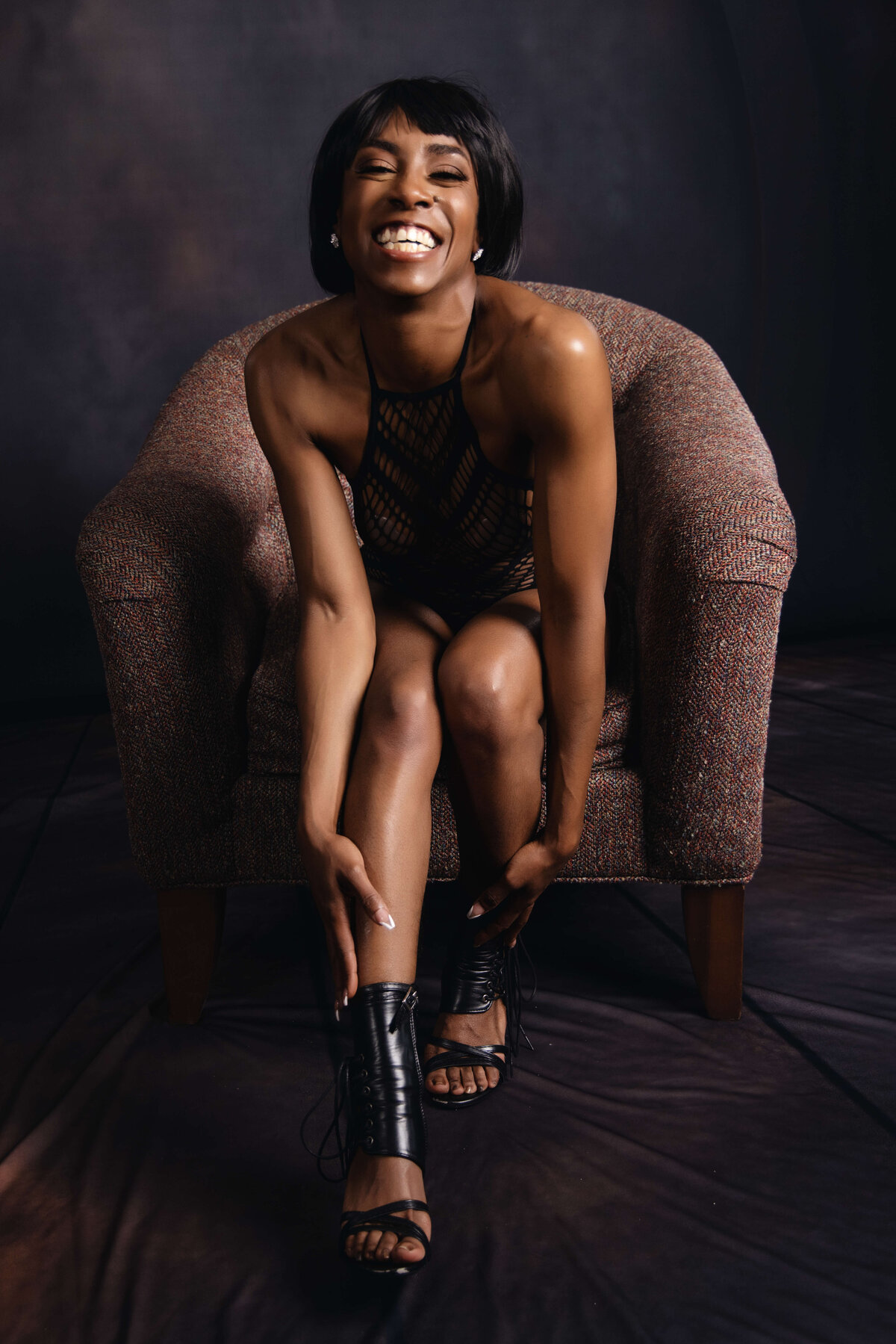 A woman with a broad smile sits on a cushioned armchair in a dark room, leaning forward to adjust her black high-heeled shoe. She wears a sleeveless black outfit and has short hair. The background is dark and blurred.