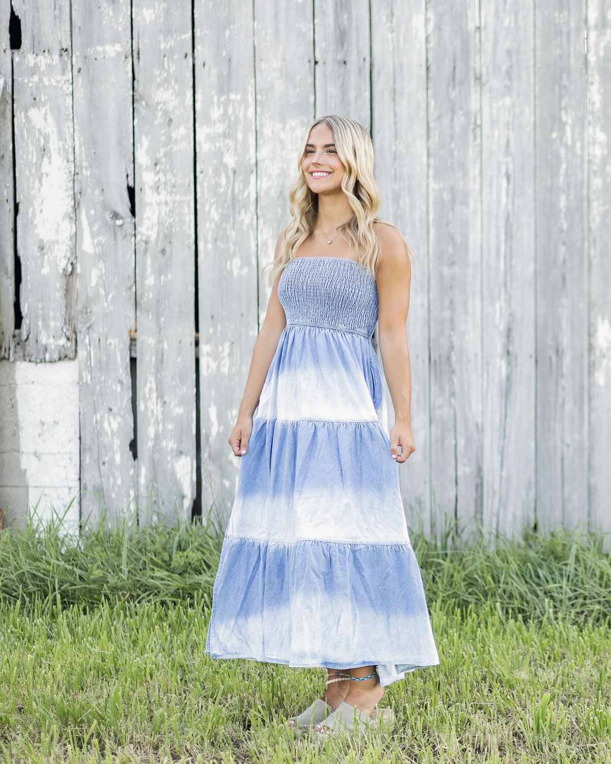 High school senior female with long blonde hair, wearing a blue and white long dress, standing in the grass, with a gray and white barn in the background, pinching the sides of her dress, looking down and smiling