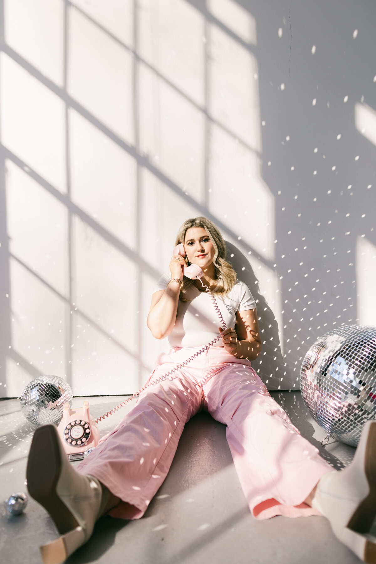 Creative branding photo featuring a woman in pink pants and a white tee sitting on the floor with a retro phone and disco balls under dappled light — captured by Dani Lacey in Greenville SC.