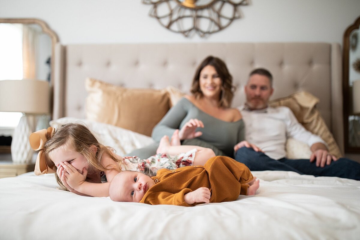 Baby on bed with big sister and parents in background by Orlando Newborn Photographer