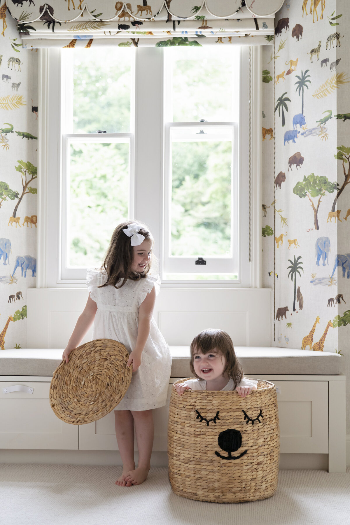 Sisters playing in their nursery. The younger girl is hiding inside a bear storage basket.