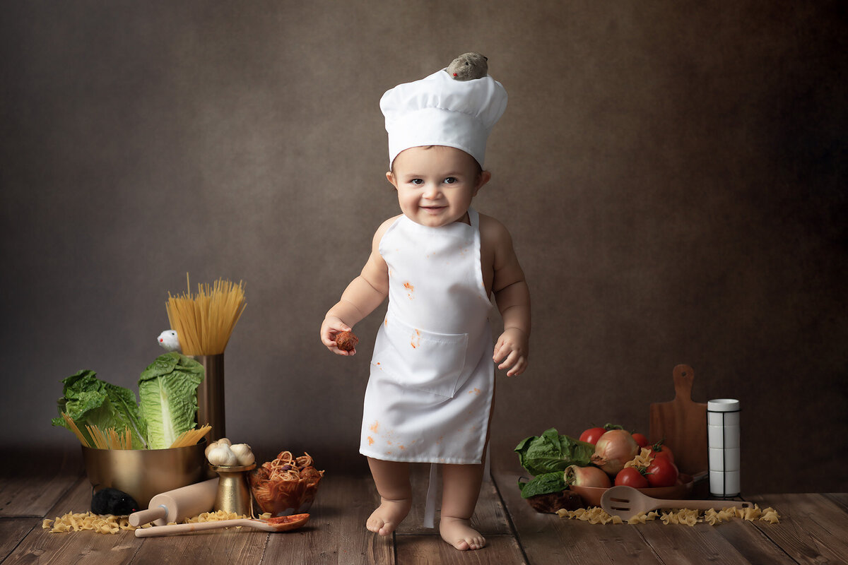 A baby dressed as a tiny chef, complete with a white apron and chef's hat, stands confidently in a kitchen-themed setup with spaghetti, fresh vegetables, and pasta scattered around, showcasing a playful Dallas milestone photography session.