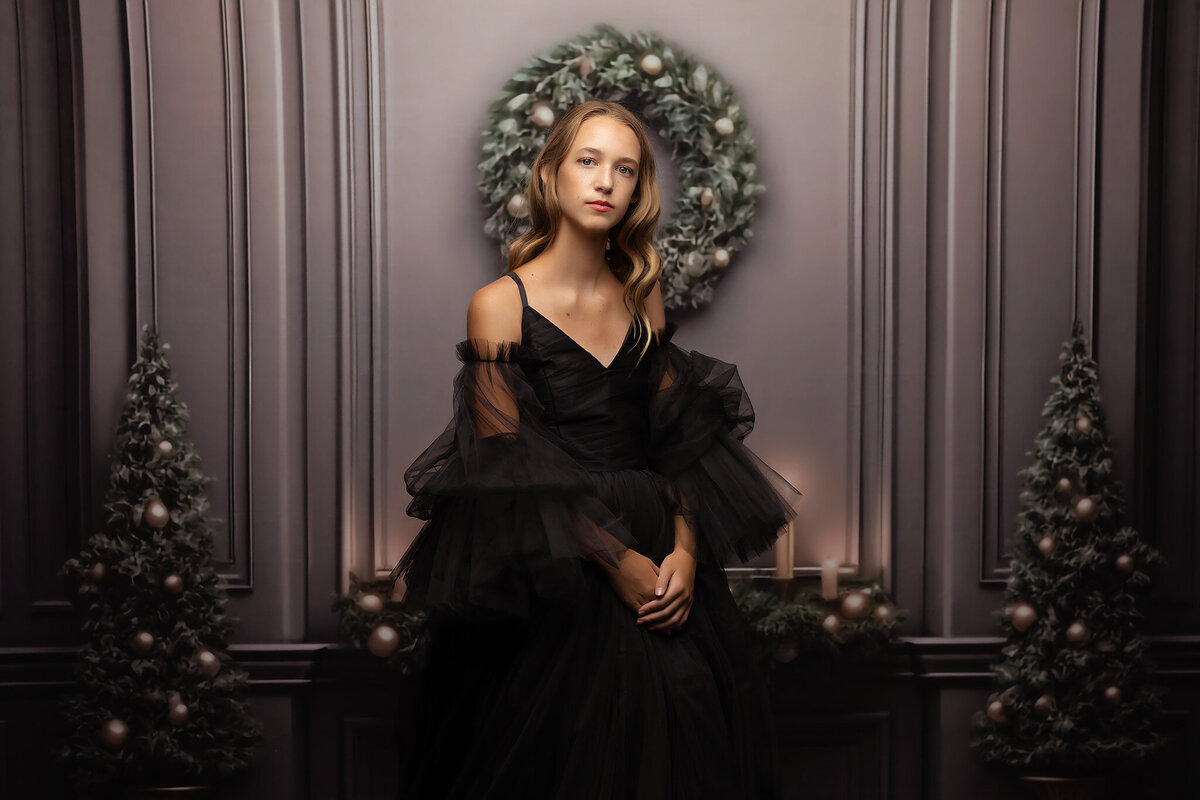 A portrait of a young woman in a flowing black gown with tulle sleeves, posed against a holiday-themed backdrop featuring a wreath and Christmas trees. The image captures a soft, ethereal look with festive touches of greenery and candles.