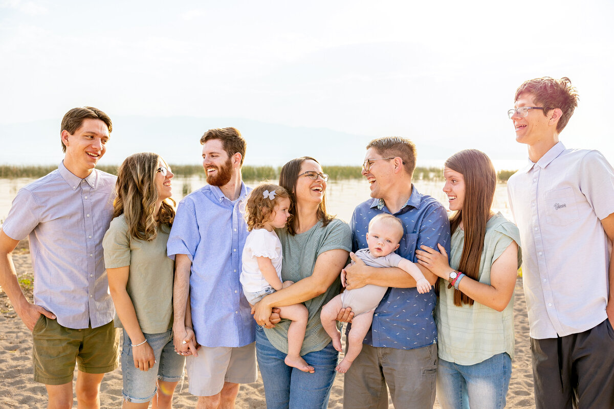 A joyful family moment as members share a laugh on the beach. A mother holds her young daughter while the father cradles their baby, surrounded by siblings and parents in the warm sunset glow.