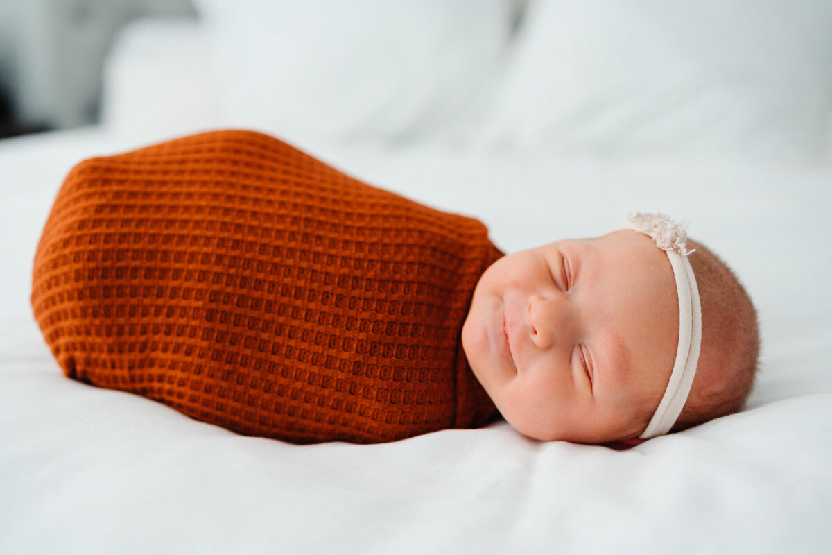 Maternity photo inspiration in Albuquerque with this charming image of a newborn baby girl. Wrapped in an orange blanket on a white mattress, the baby is smiling contentedly with her eyes closed, showcasing a peaceful and joyful moment.