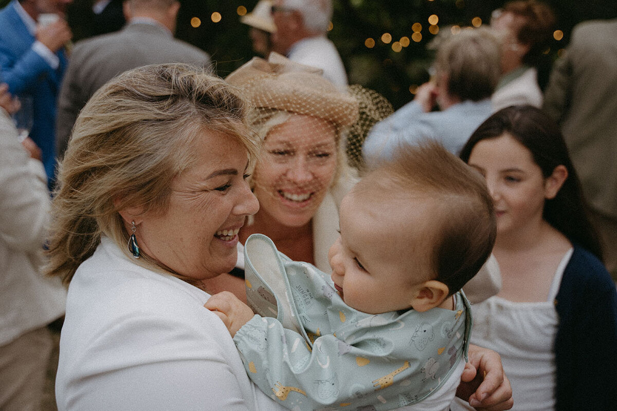 Gwladys sourit en regardant une invitée portant un bébé. Une scène tendre capturée par Kamescop, photographe et vidéaste en Bretagne.