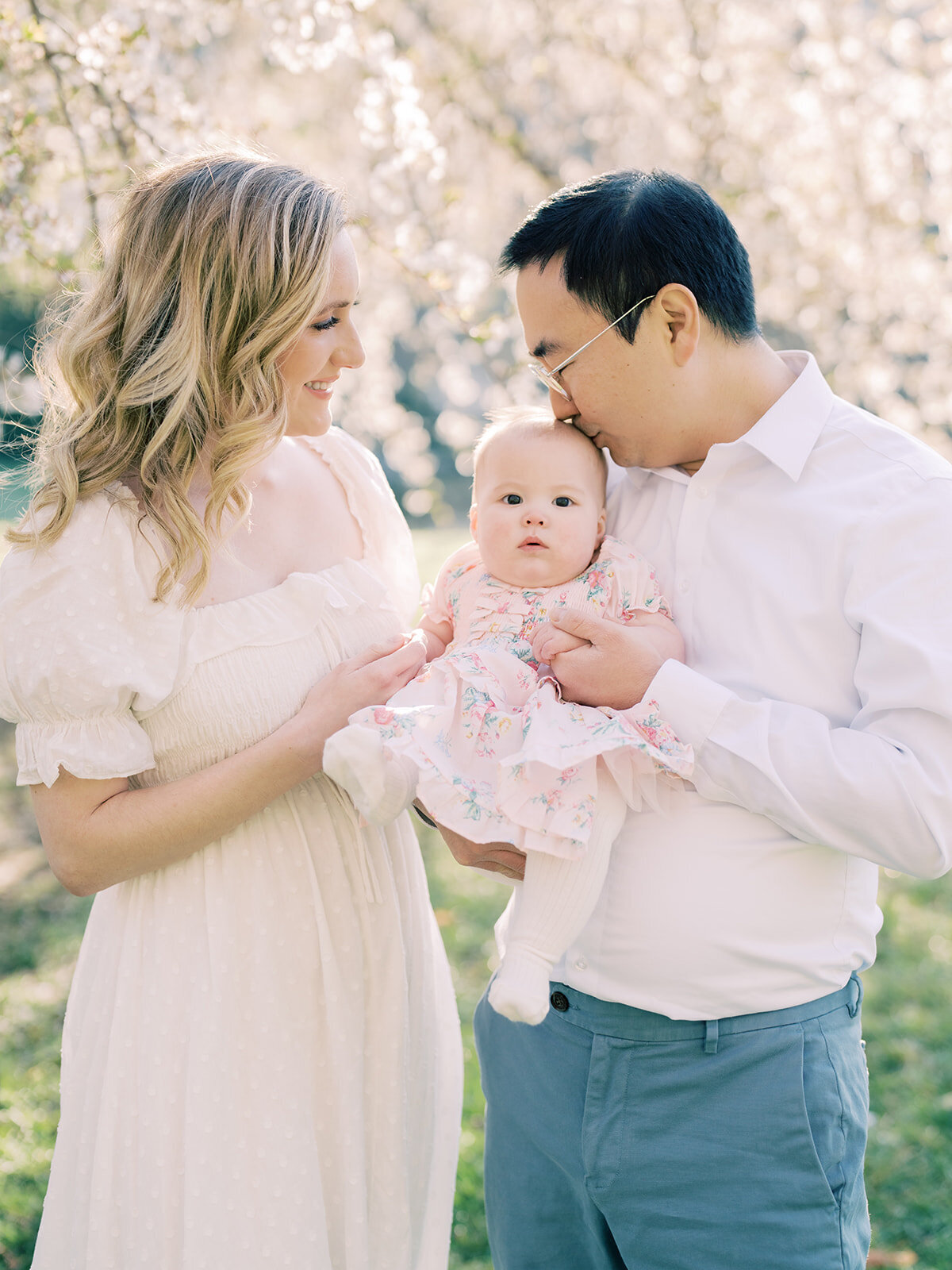 Blonde mother and Asian father hold their little girl in front of cherry blossom tree as father kisses baby's head.