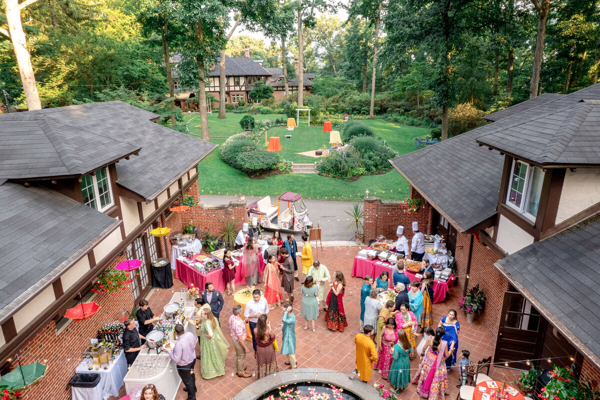 A vibrant outdoor celebration at a large residential property. Guests in colorful traditional attire gather around tables with food in a courtyard. The setting features lush greenery and quaint buildings, suggesting a festive, multicultural event.