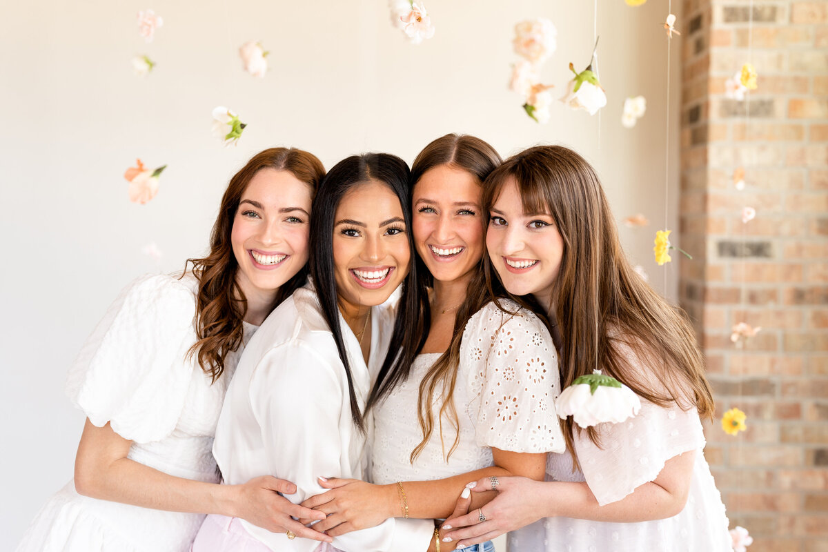 Texas A&M senior girls hugging and laughing surrounded by hanging flowers at Bravely Studio