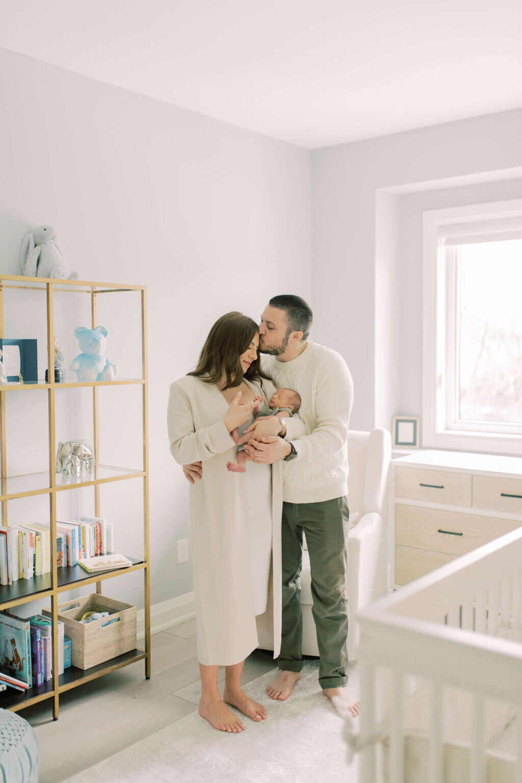 Couple standing in nursery holding their newborn, father kissing mother’s temple – peaceful family connection