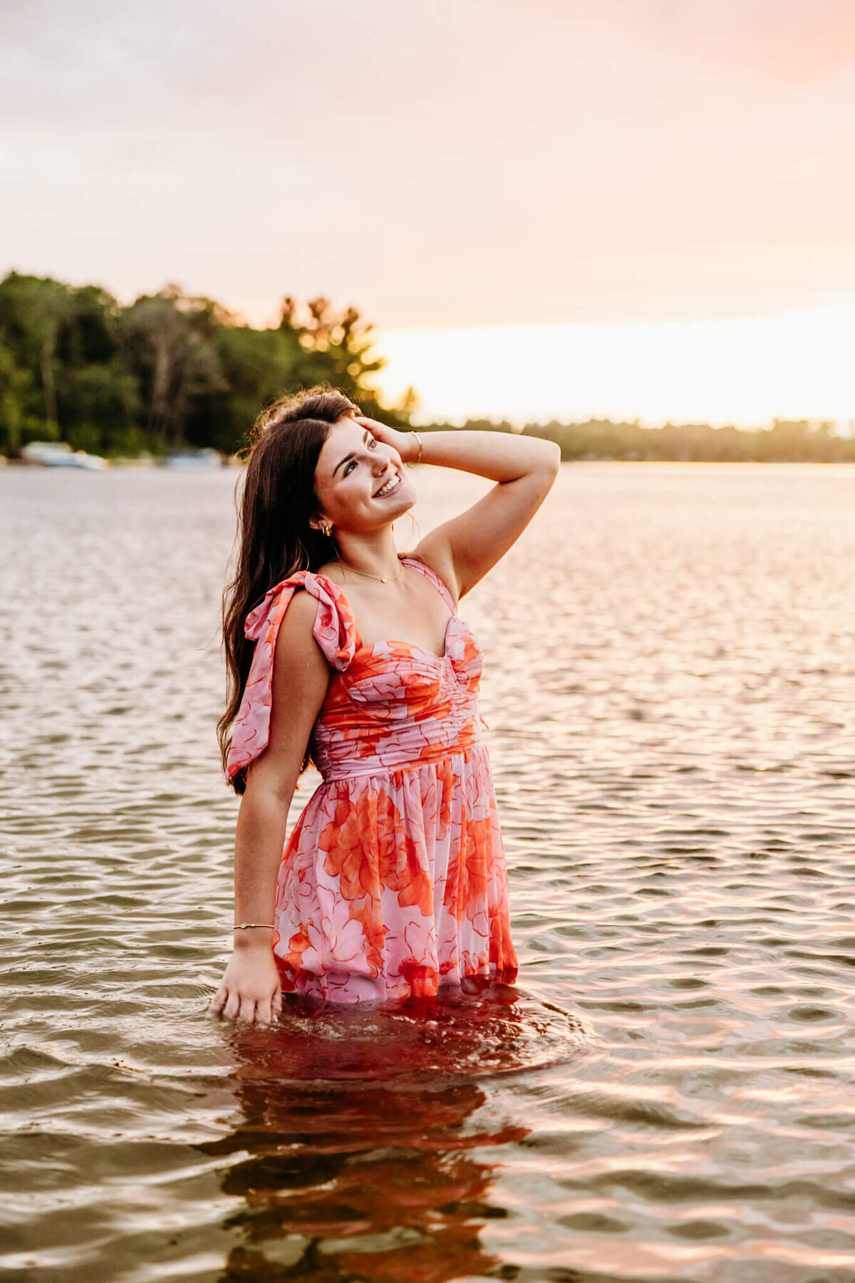 teenage girl in a pink and orange patterned dress standing in a lake and playing with her hair as she touches the water for her Green Bay senior photography session.