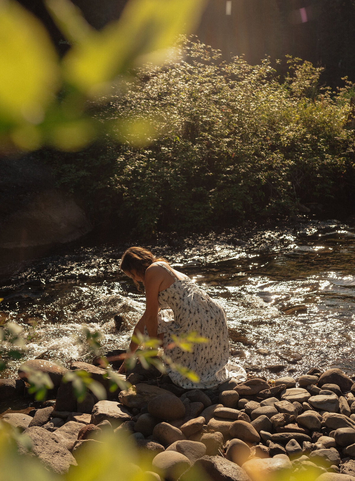 woman in white dress sits near the river and observes the river rocks on a sunny warm day in Bend, Oregon