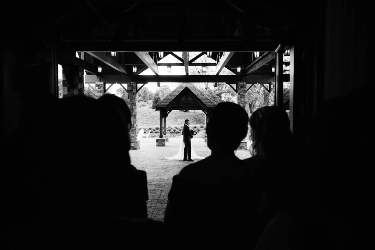 A silhouetted couple stands facing each other under a wooden gazebo during a wedding ceremony, beautifully captured by an NJ wedding photographer, while the outlines of seated guests are visible in the foreground.