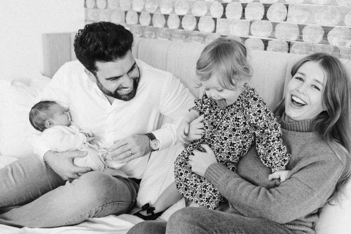 Parents sitting on a couch with their two young children, laughing as the toddler sits between them.