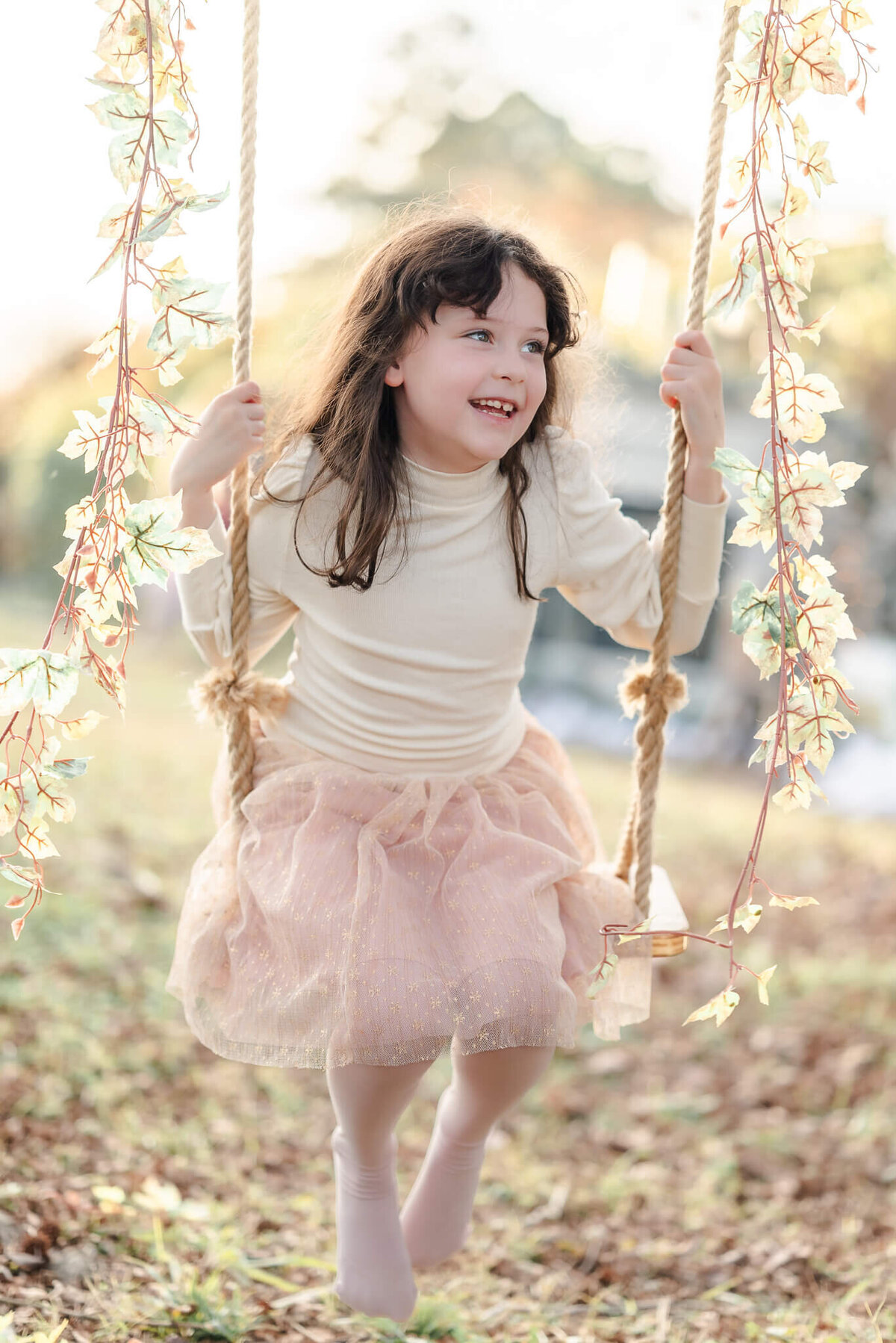 A young girl, wearing pink and cream smiles as she swings on a wooden swing.