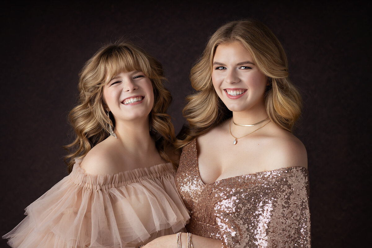 Two young women share a joyful moment, dressed in a soft, off-the-shoulder tulle gown and a sparkling rose gold sequin dress. Their smiles shine against a dark, textured background, making this a perfect example of luxury Dallas senior photography.